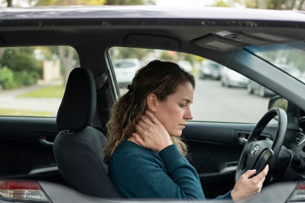 Woman sits in the driver's seat of a car, looking at her smartphone while resting or rubbing her neck with her hand.